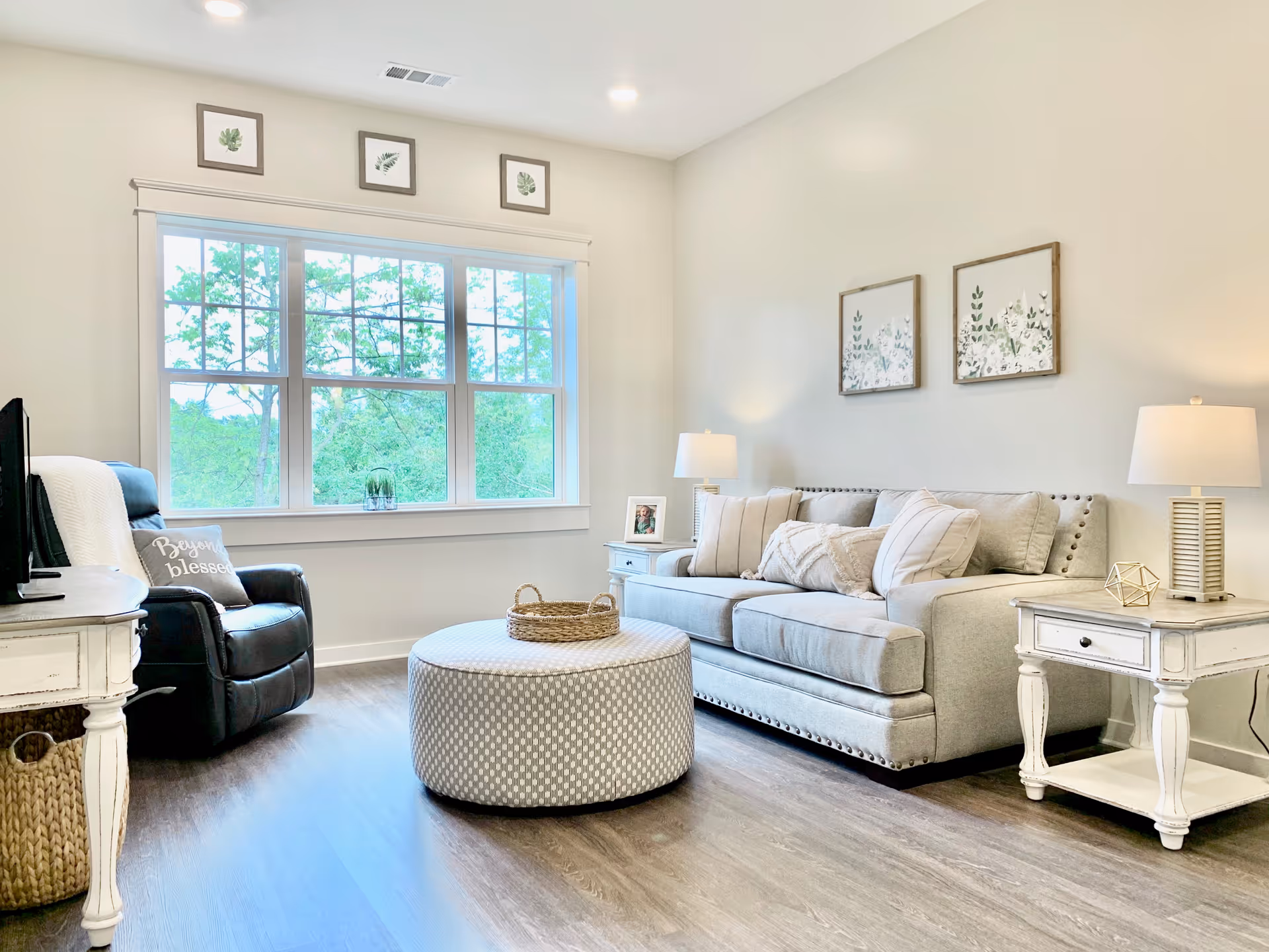 Bright living room with a gray sofa, armchair, round ottoman, side tables, lamps, and a large window overlooking trees.
