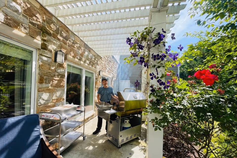 A man standing next to a stainless steel grill on a patio with a stone wall and large windows on one side. The patio is covered with a white pergola, and there are purple flowers climbing a white post. On the other side, there are green bushes and red flowers under a bright blue sky.