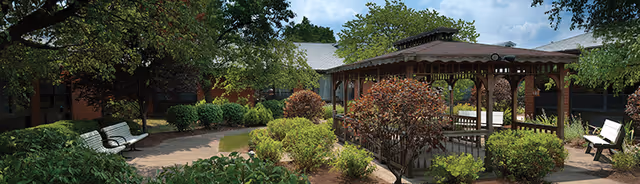 A landscaped outdoor garden area at Cranberry Place featuring a wooden gazebo surrounded by various green shrubs and trees, with benches placed along a paved pathway under a partly cloudy sky.