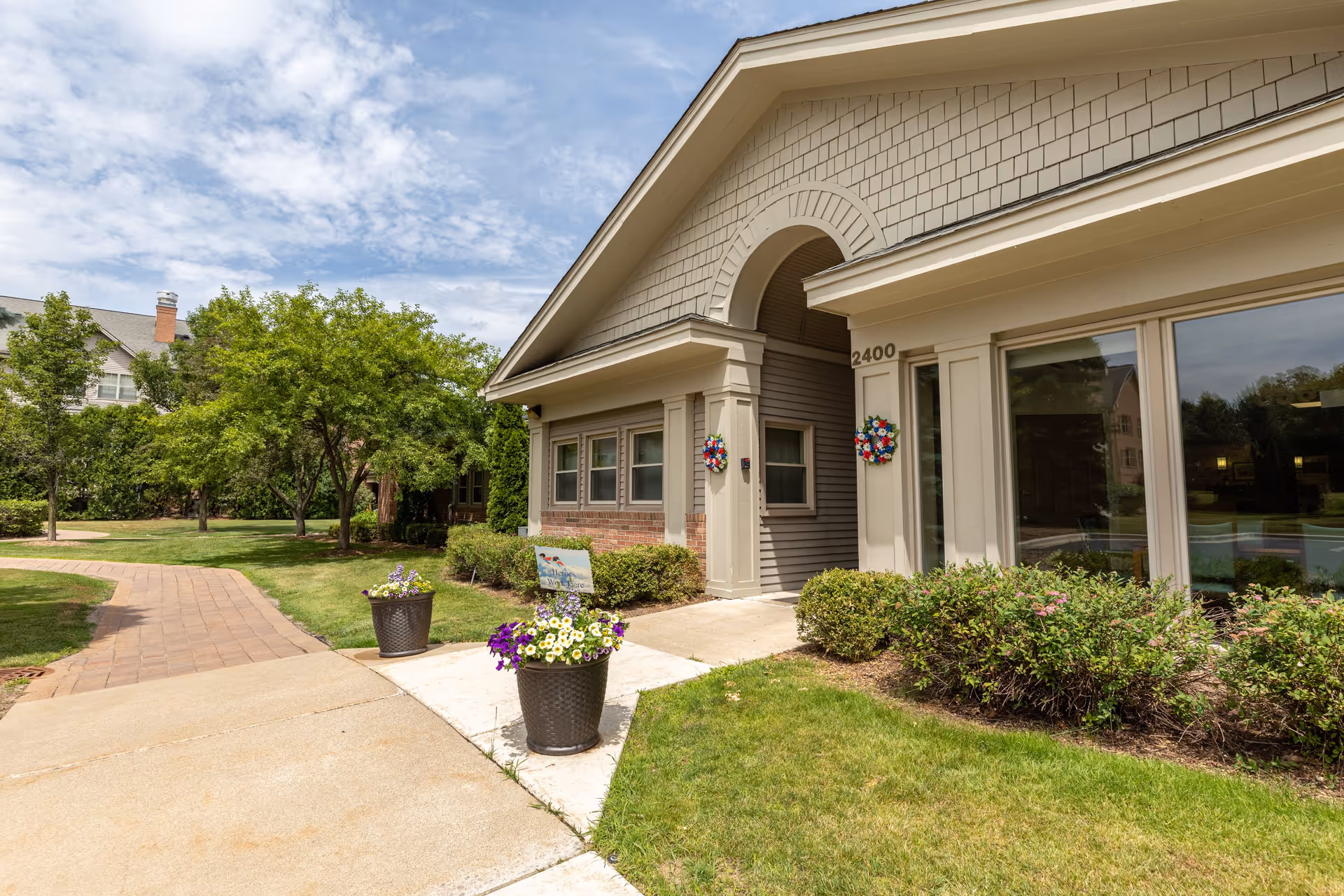 Exterior view of a senior living facility building with beige siding and brick accents. The building has large windows and two decorative wreaths on the walls near the entrance. The address number 2400 is visible. There are two large flower pots with colorful flowers near the sidewalk, and a paved walkway curves through a well-maintained grassy area with trees and shrubs under a partly cloudy sky.