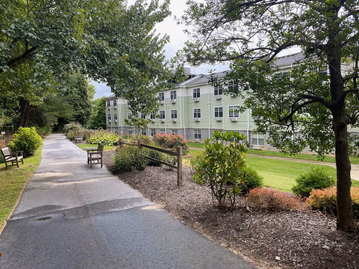 Paved walkway with benches and landscaped gardens alongside a pale green multi-story retirement building under trees.