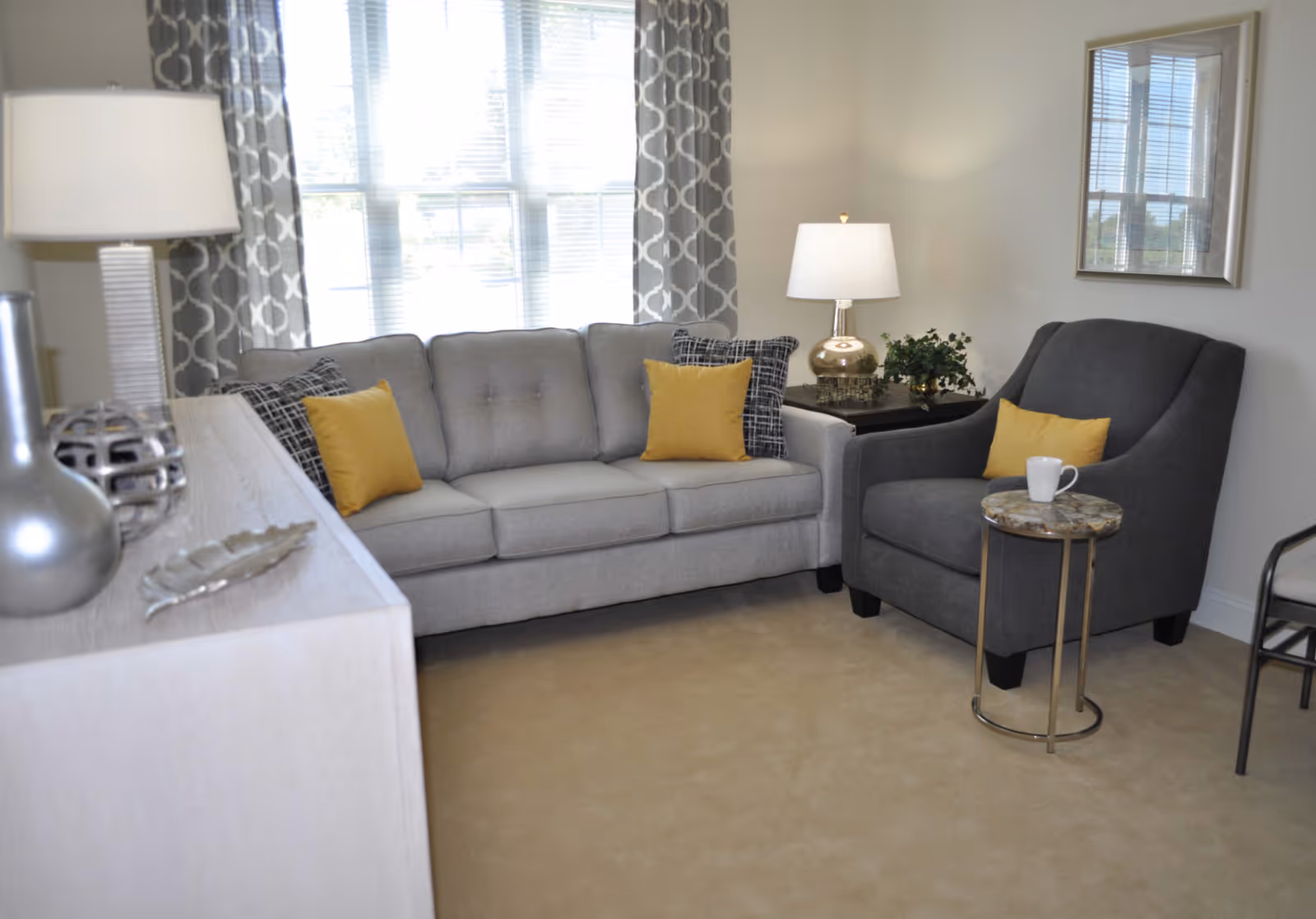 Bright living room with a gray sofa and matching armchair accented by yellow pillows, side tables, and lamps.