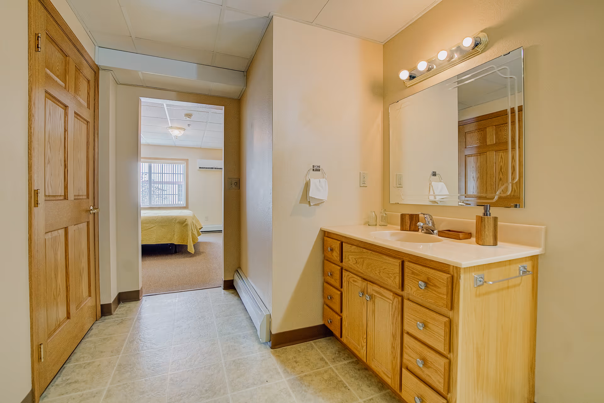 Interior view of a bathroom area with a wooden vanity, sink, mirror, and light fixtures above. To the left, there is a doorway leading to a bedroom with a bed covered in a yellow blanket and a window with vertical blinds.