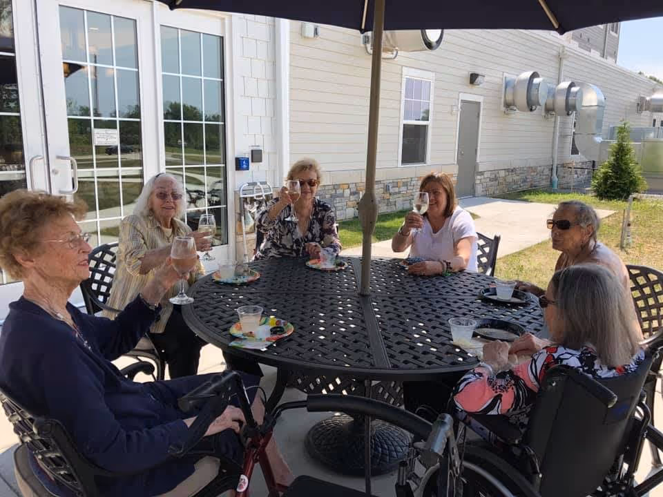Six elderly women sitting around a round outdoor metal table under a large umbrella, raising glasses in a toast. They are outside near a building with large windows and stone accents, enjoying a sunny day.