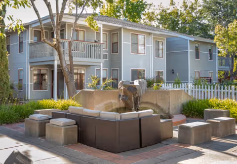 Outdoor seating area with cushioned wicker chairs and concrete stools arranged around a water fountain, set in front of a two-story gray building with white trim and a balcony, surrounded by trees and greenery.
