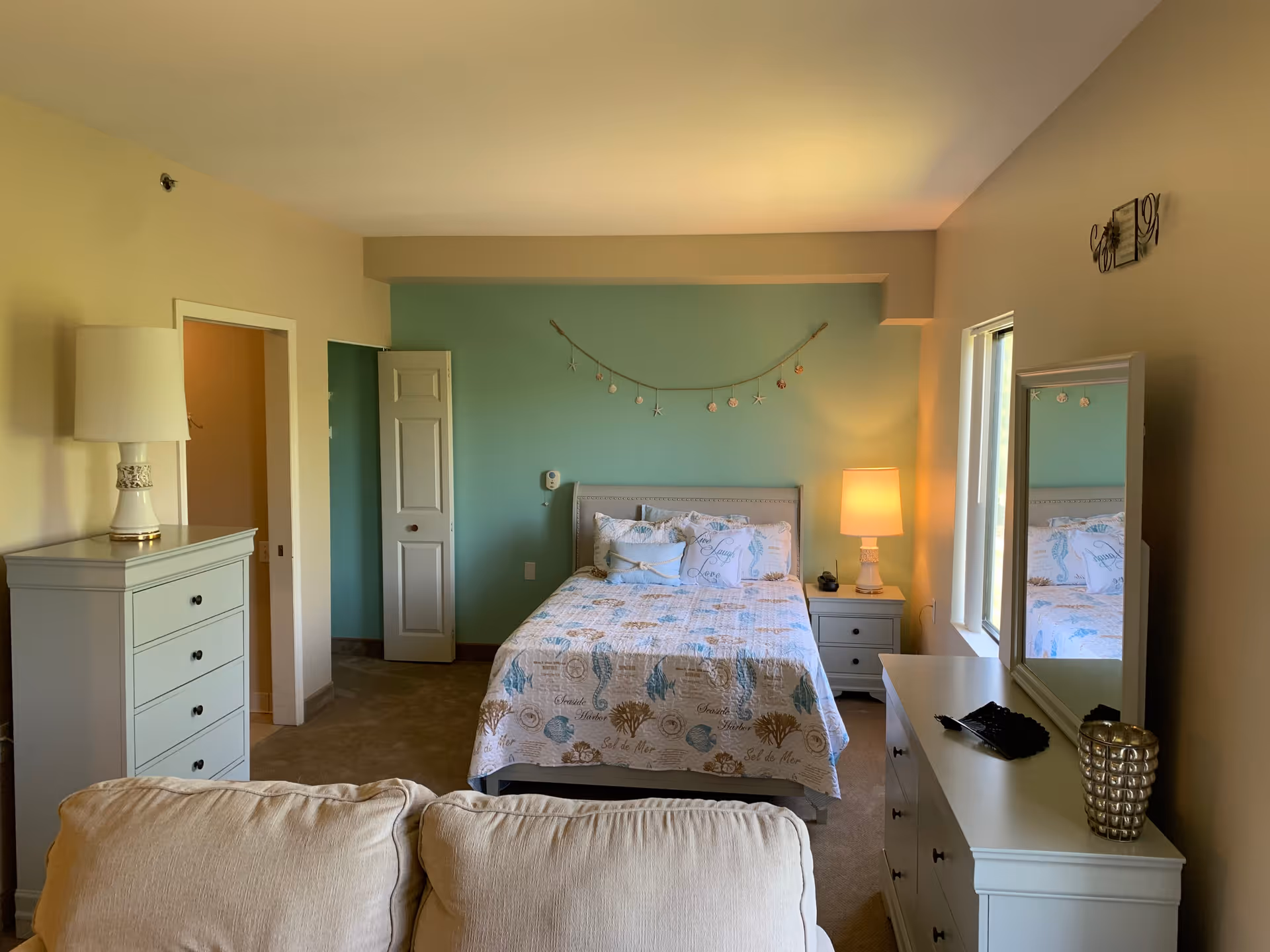 A cozy bedroom with a bed centered against a teal accent wall, decorated with a seashell and starfish garland. The bed has a quilt with sea-themed patterns and several pillows. To the right of the bed is a nightstand with a lamp and a phone. A dresser with a mirror is positioned under a window on the right side of the room. On the left side, there is a tall chest of drawers with a lamp on top. The room has beige walls and carpet, with a beige couch partially visible in the foreground.