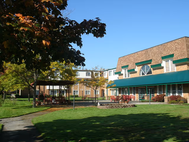 Outdoor courtyard with lawn, gazebo, patio seating, and a brick senior living building with green awnings under a clear blue sky.