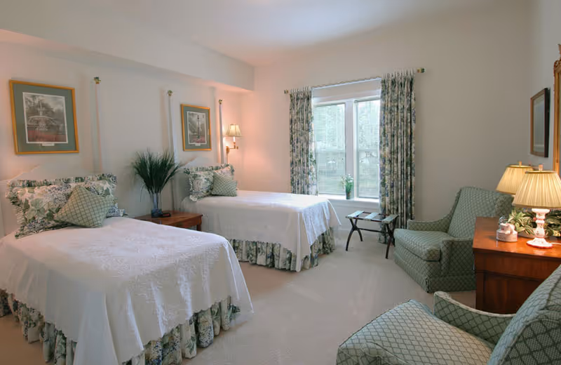 A bright and cozy bedroom with two twin beds dressed in white bedspreads and floral bed skirts. Each bed has multiple pillows with floral and patterned designs. Between the beds is a wooden nightstand with a potted plant and a wall-mounted lamp above it. The room features a window with floral curtains letting in natural light, a small table with a plant, and two upholstered armchairs with a diamond pattern. A wooden dresser with a lamp and decorative items is also visible.