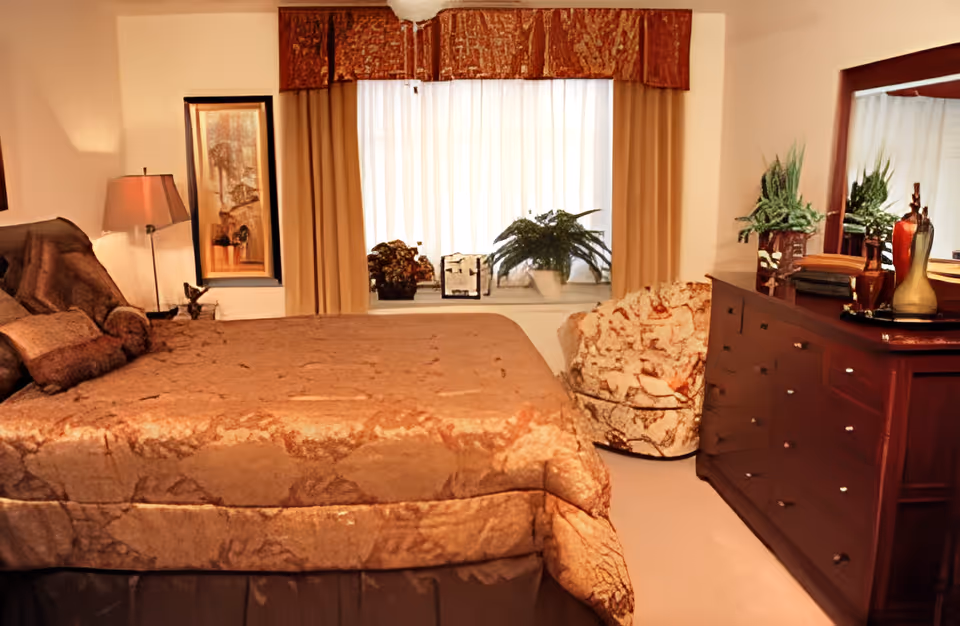 A cozy bedroom with a large bed covered in a patterned brown bedspread, a cushioned armchair with a floral design, a wooden dresser with a large mirror, and two potted plants on the windowsill behind beige curtains.