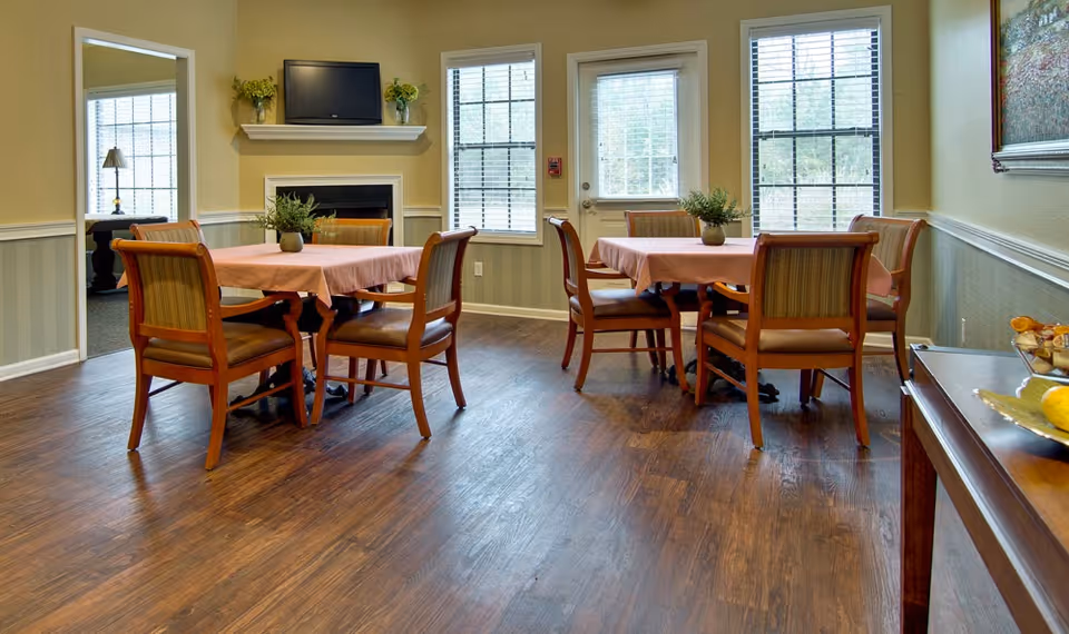 A cozy dining room with two square tables covered with pink tablecloths, each surrounded by four wooden chairs with cushioned seats. The room has wooden flooring, beige walls with white wainscoting, and three large windows letting in natural light. A small TV is mounted above a fireplace, and there are plants on the tables and mantel. A door with a window is visible between the windows.