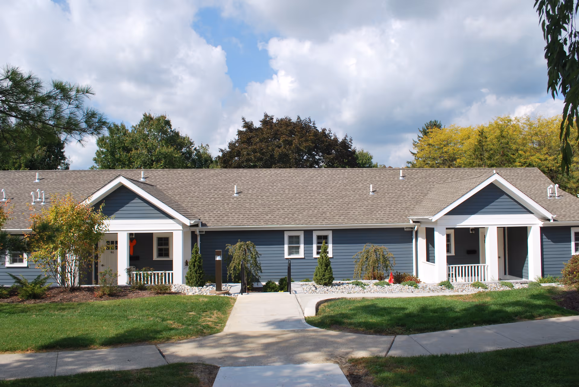 Exterior view of a single-story blue building with white trim, featuring two covered porch entrances, surrounded by green grass, small trees, and shrubs under a partly cloudy sky.