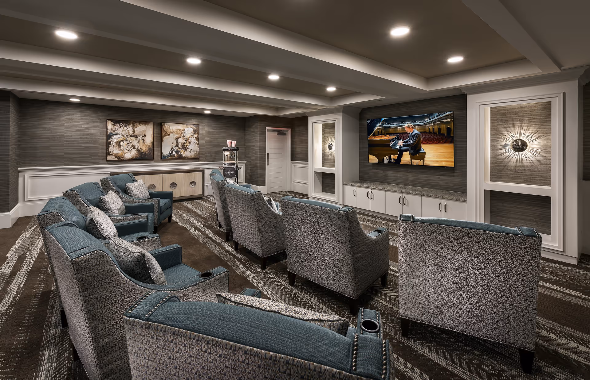 A cozy media room with two rows of upholstered armchairs facing a large flat-screen TV mounted on the wall. The TV displays a man playing a grand piano on a stage. The room features modern recessed lighting, patterned carpet, decorative wall art, and a water dispenser in the corner.
