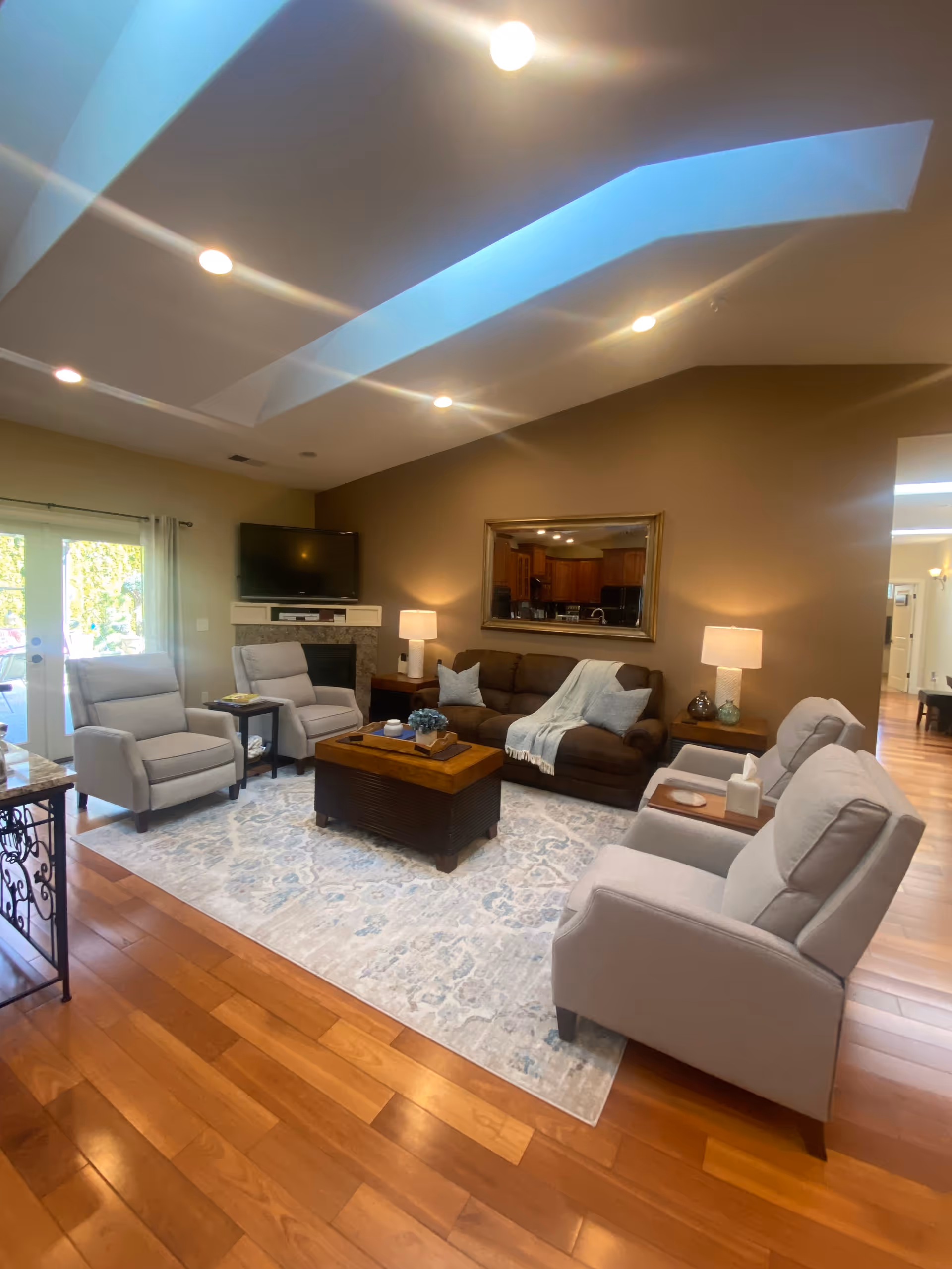 A cozy living room with wooden flooring, a large area rug, a brown sofa with a throw blanket, four light gray armchairs, two side tables with lamps, a coffee table with decorative items, a wall-mounted TV above a fireplace, and a large mirror reflecting the kitchen area.