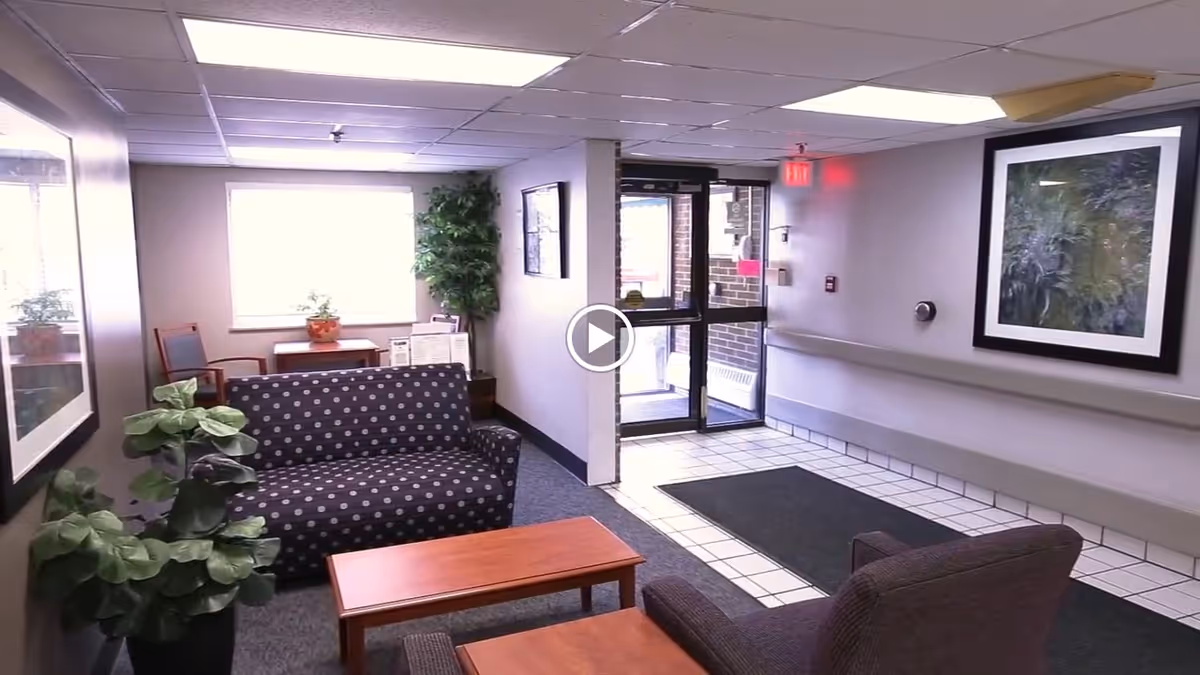 A small waiting area inside a facility with a patterned couch, an armchair, a wooden coffee table, potted plants, framed artwork on the walls, and a glass door leading outside.