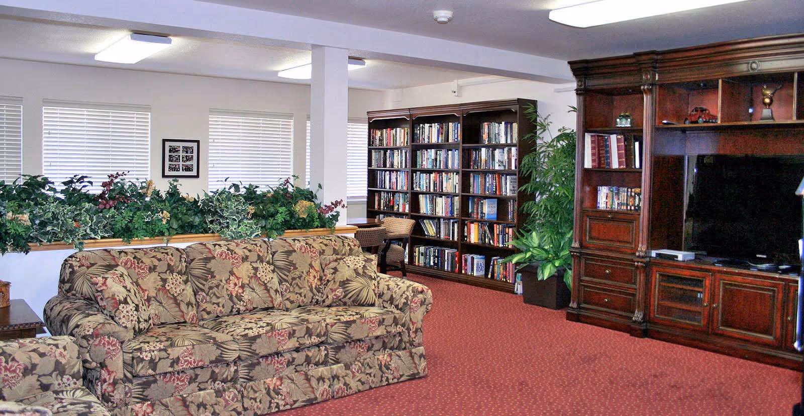 A senior living community common room with floral upholstered sofas, bookshelves, a large wooden entertainment center, and potted plants.