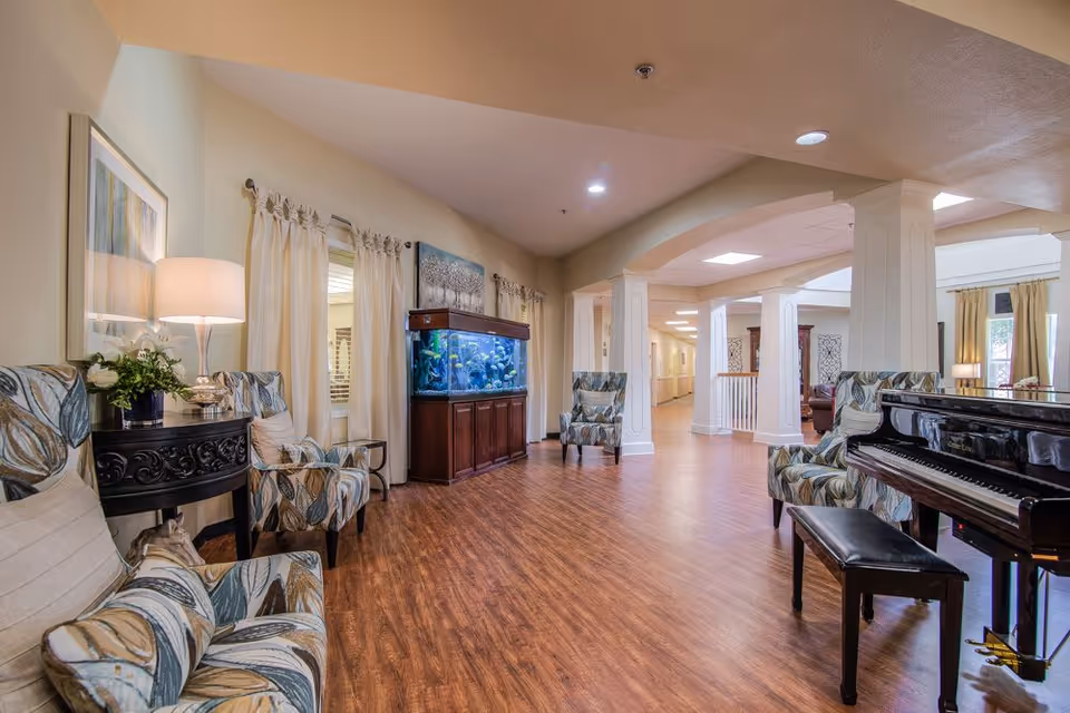 A spacious senior living facility common area with wooden flooring, patterned armchairs, a large fish tank, a black grand piano with a bench, and soft lighting from lamps and ceiling lights. The room has cream-colored walls and curtains, with decorative artwork on the walls and white columns supporting the ceiling.
