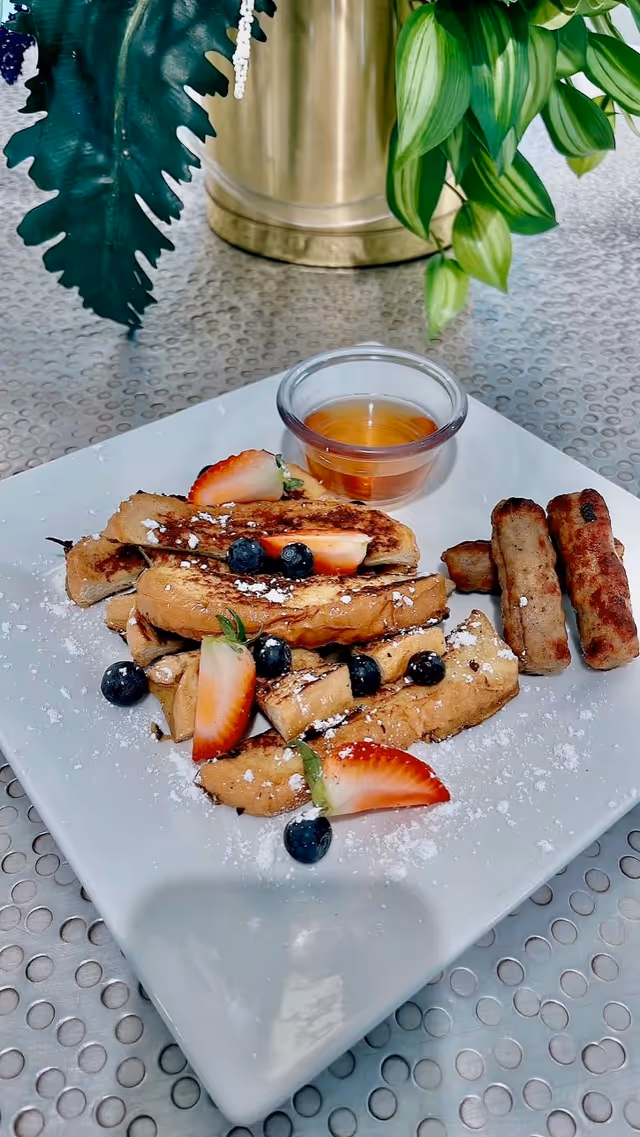 A white square plate with French toast sticks topped with powdered sugar, fresh blueberries, and sliced strawberries, served with a small cup of syrup and three sausage links on the side. The plate is on a textured metal table with green leafy plants in the background.