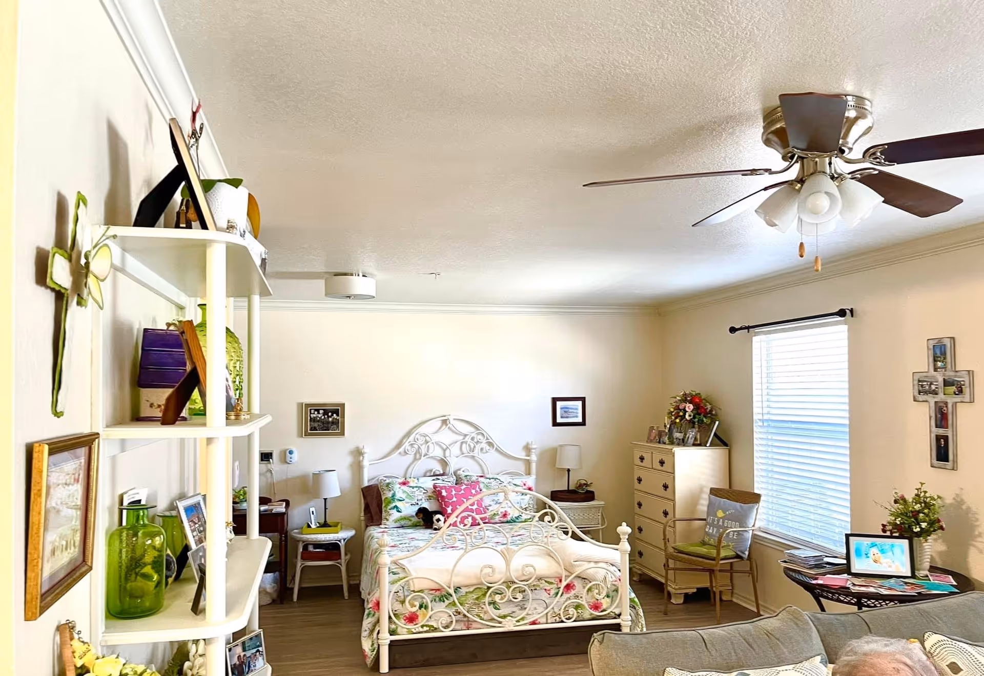A cozy bedroom with a white ornate metal bed frame, floral bedding, and decorative pillows. The room features a ceiling fan with lights, a window with blinds, a wooden dresser with flowers on top, a small round table with a digital photo frame, and a chair with a cushion. There are shelves on the left wall holding various decorative items and framed pictures, and the walls are adorned with crosses and framed artwork.