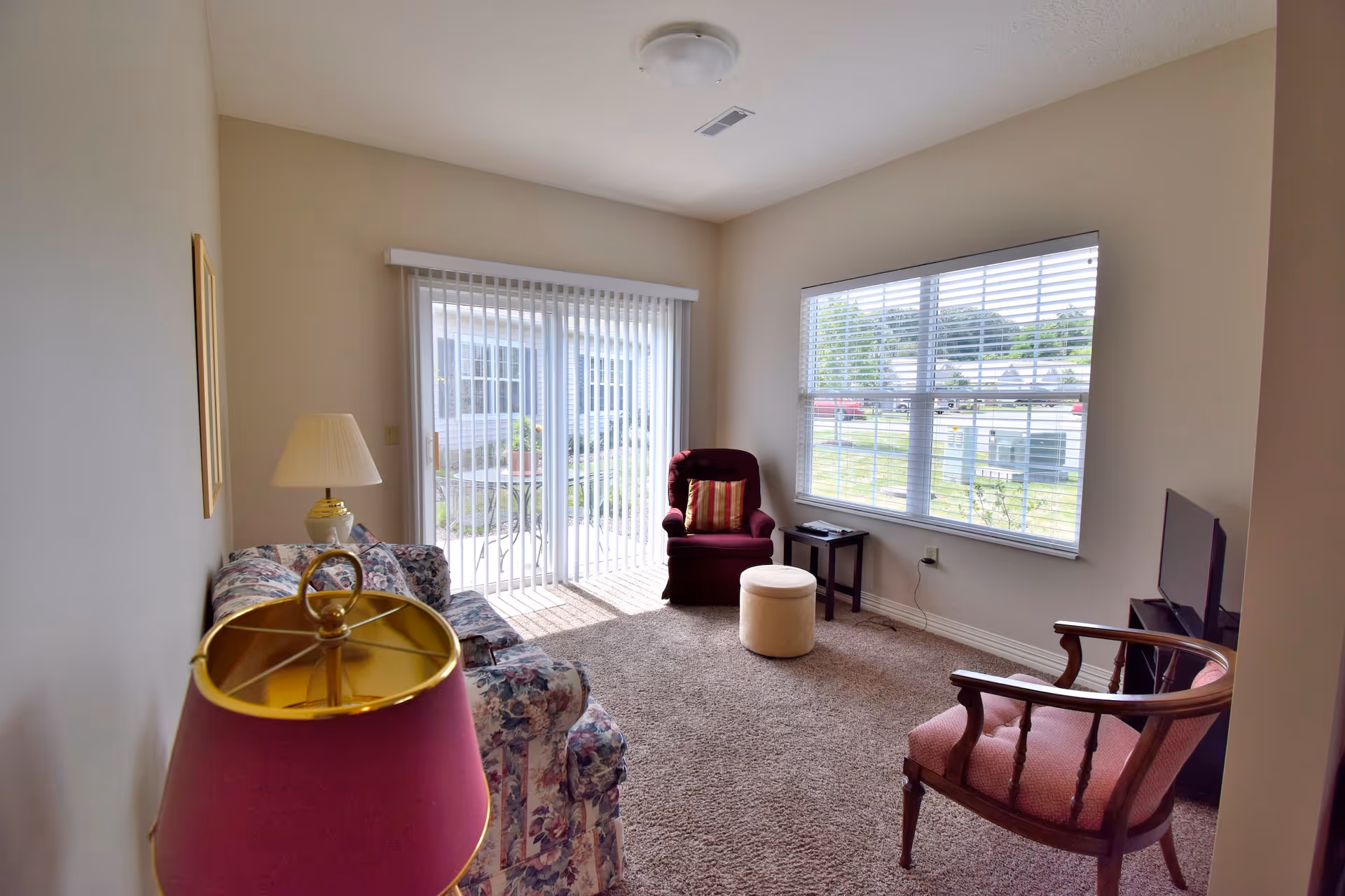Carpeted living room with a floral sofa, two armchairs, lamps, a TV, a sliding glass door and a large window letting in daylight.