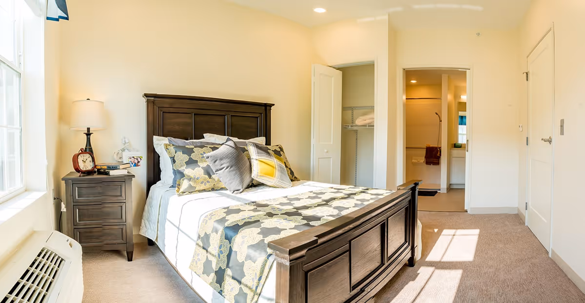 A bright bedroom with a large wooden bed featuring a patterned bedspread and multiple pillows. To the left of the bed is a wooden nightstand with a lamp, clock, and some books. There is a window on the left wall letting in natural light. The room has beige walls and carpeted floor. In the background, there is an open closet door and a doorway leading to a bathroom.