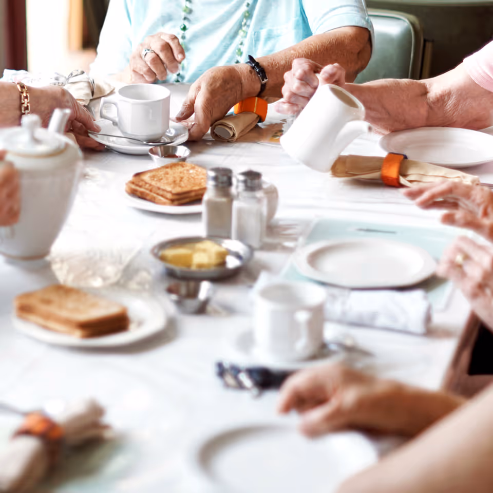 Close-up of elderly individuals sitting around a table with white tablecloth, having tea or coffee. The table has plates with toast, butter, salt and pepper shakers, napkins with orange rings, and white cups and saucers.