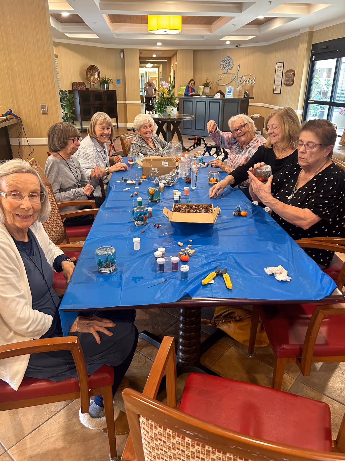A group of elderly women sitting around a table covered with a blue plastic tablecloth, engaging in a craft activity with various small containers, decorations, and tools. They are smiling and appear to be enjoying the activity in a well-lit room with beige walls and a reception desk in the background with the name 'Atria' displayed on the wall.