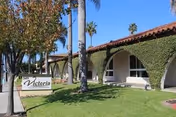 Exterior view of Victoria Healthcare and Rehabilitation facility with a lawn, palm trees, and a building covered partially with ivy under a clear blue sky.