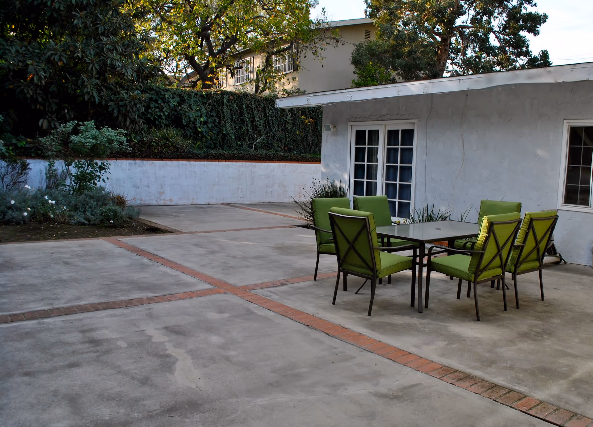 Outdoor patio area with a concrete floor featuring brick accents, a table with six green cushioned chairs, and a white building with French doors and a window. There is greenery and trees in the background.