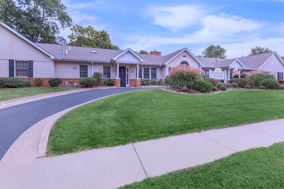 Exterior view of Country Manor Memory Care facility showing a single-story building with beige walls, a dark roof, and a curved driveway leading to the entrance. The front yard is well-maintained with green grass, bushes, and ornamental grasses. A sign near the entrance reads 'Country Manor Memory Care'.