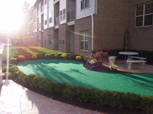 Outdoor area of a senior living facility with a putting green surrounded by neatly trimmed bushes and yellow flowers. There is a concrete pathway and a round stone table with benches near the building. The building has brick and siding exterior with multiple windows and balconies.