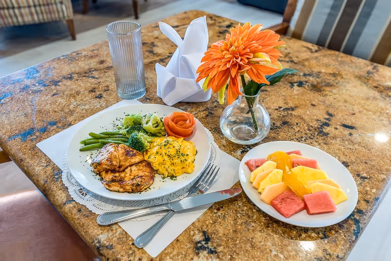 A meal served on a marble table with a plate containing grilled chicken, scrambled eggs, green beans, broccoli, and a tomato rose garnish. Next to it is a plate with assorted fresh fruit including watermelon, pineapple, and cantaloupe. A glass of water and a small vase with an orange flower are also on the table, along with a folded white napkin.