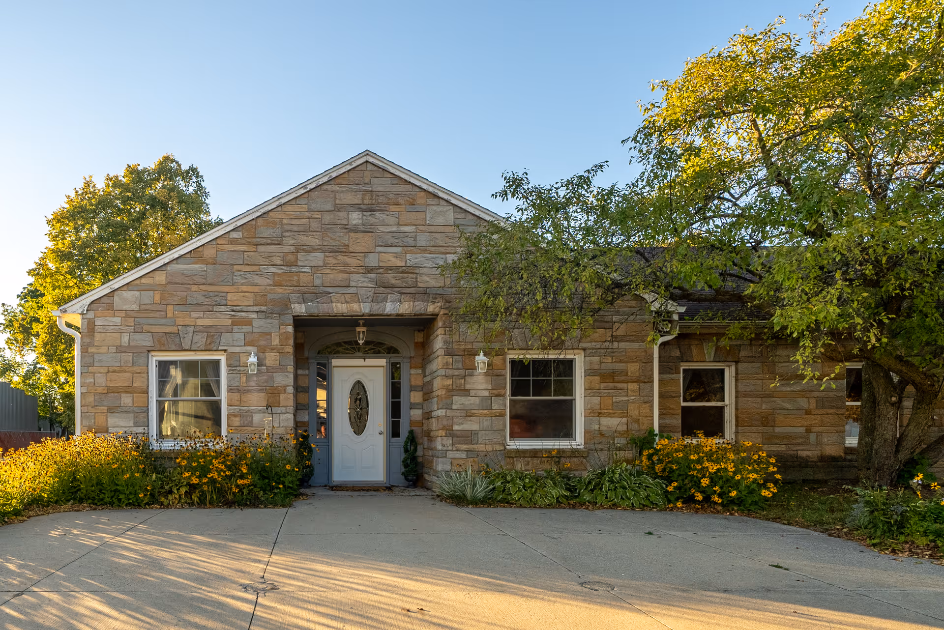 Front view of a single-story stone building with a white entrance door, windows, and surrounding landscaping.