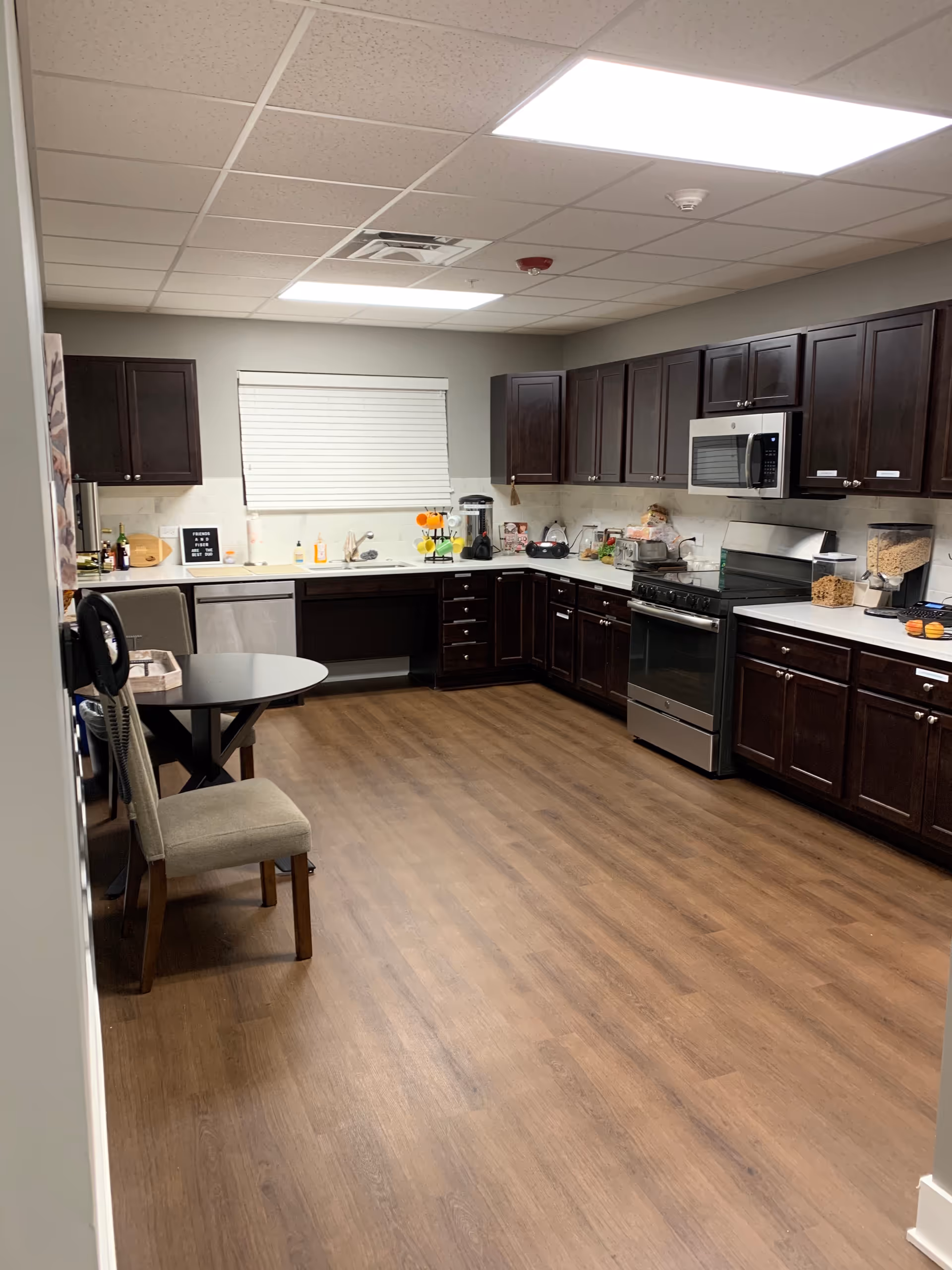 A spacious kitchen with dark wooden cabinets, a stainless steel stove and microwave, a dishwasher, and a window with closed blinds. The kitchen has a light-colored countertop with various kitchen items and appliances on it. There is a small round table with two chairs on the left side and wood-look flooring.