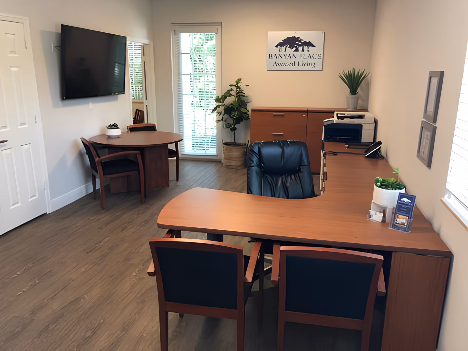An office space with a wooden desk and black leather office chair. Two wooden chairs with black cushions are placed in front of the desk. Behind the desk is a wooden cabinet with a printer and a potted plant on top. A round wooden table with two chairs is positioned near a wall-mounted flat screen TV. A door and a window with blinds are visible, along with a sign on the wall that reads 'Banyan Place Assisted Living'.
