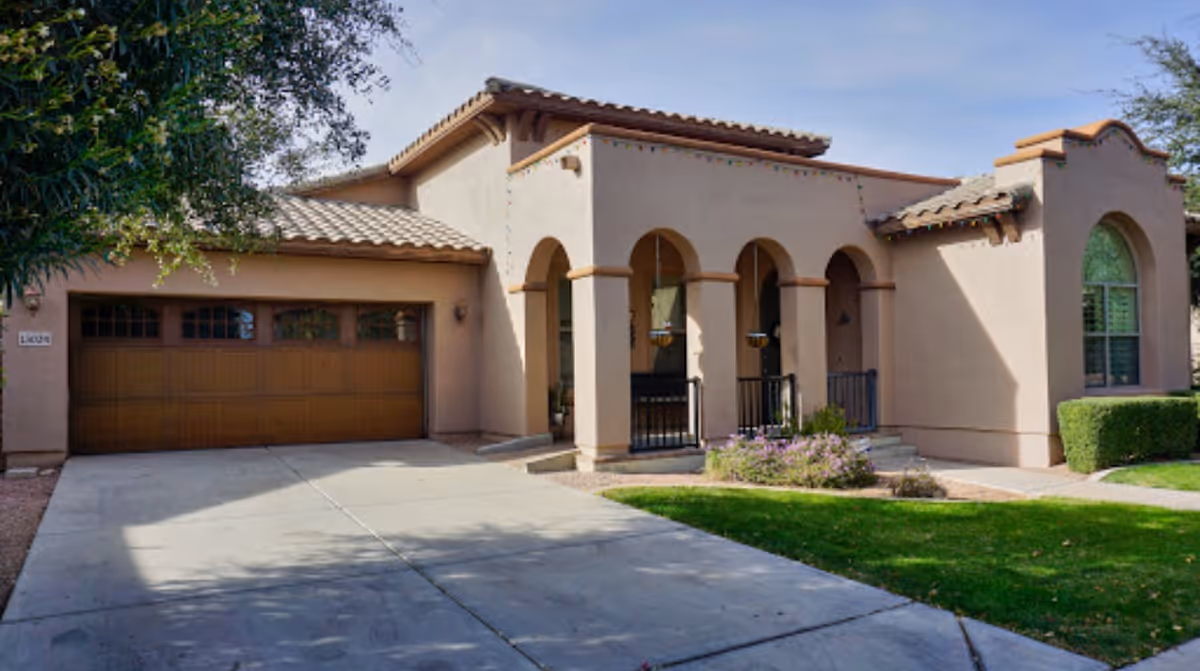Exterior view of a single-story house with a tiled roof, arched front porch, and a two-car garage. The driveway and front yard with green grass and some shrubs are visible under a clear blue sky.