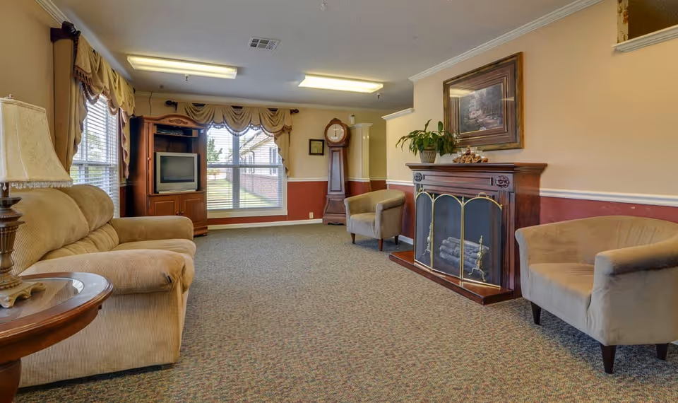 A carpeted common living room with sofas, armchairs, a fireplace, TV cabinet, and large windows.