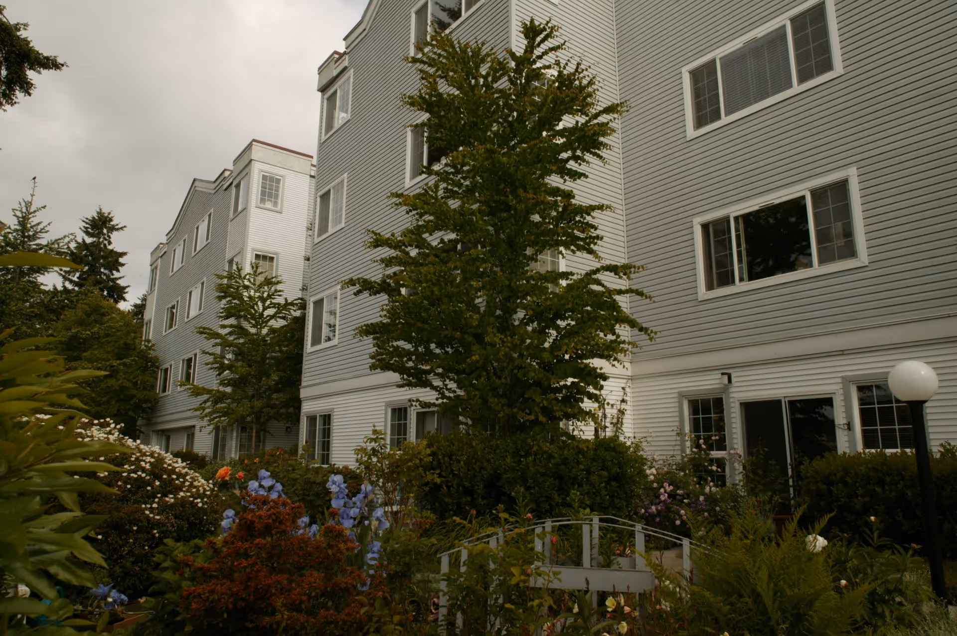 Exterior view of a multi-story senior living facility building with light gray siding, multiple windows, and a garden area in the foreground featuring various plants, flowers, and small trees under a cloudy sky.