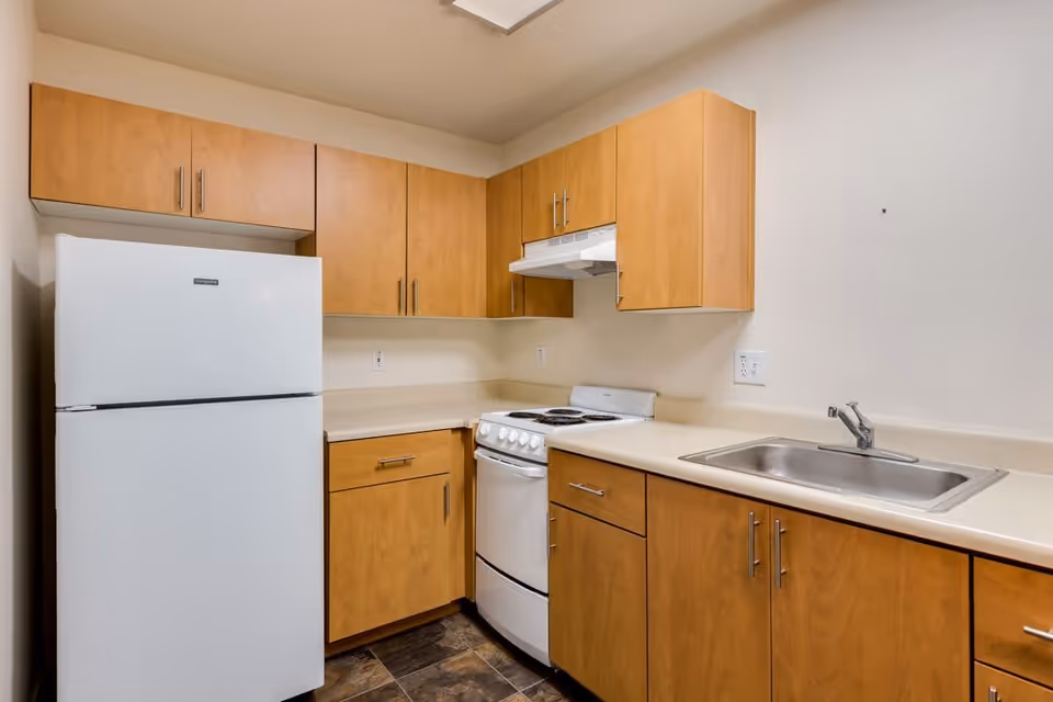 A small kitchen area with light wood cabinets, a white refrigerator, a white electric stove with an overhead vent, and a stainless steel sink set in a beige countertop. The floor has dark tile, and the walls are painted light beige.