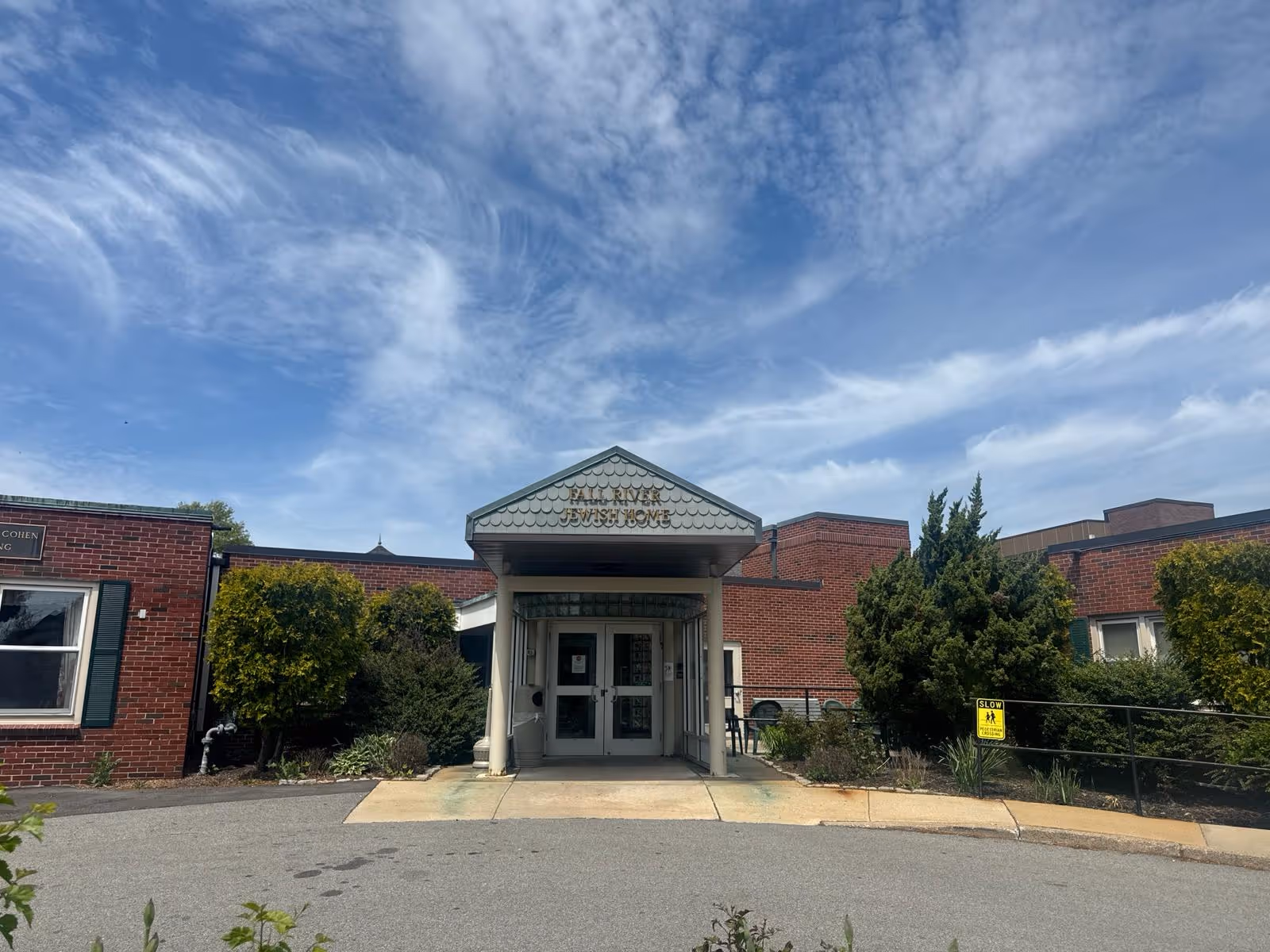 Front entrance of a single-story brick senior living facility with a covered canopy, double glass doors, and surrounding shrubs under a blue sky.
