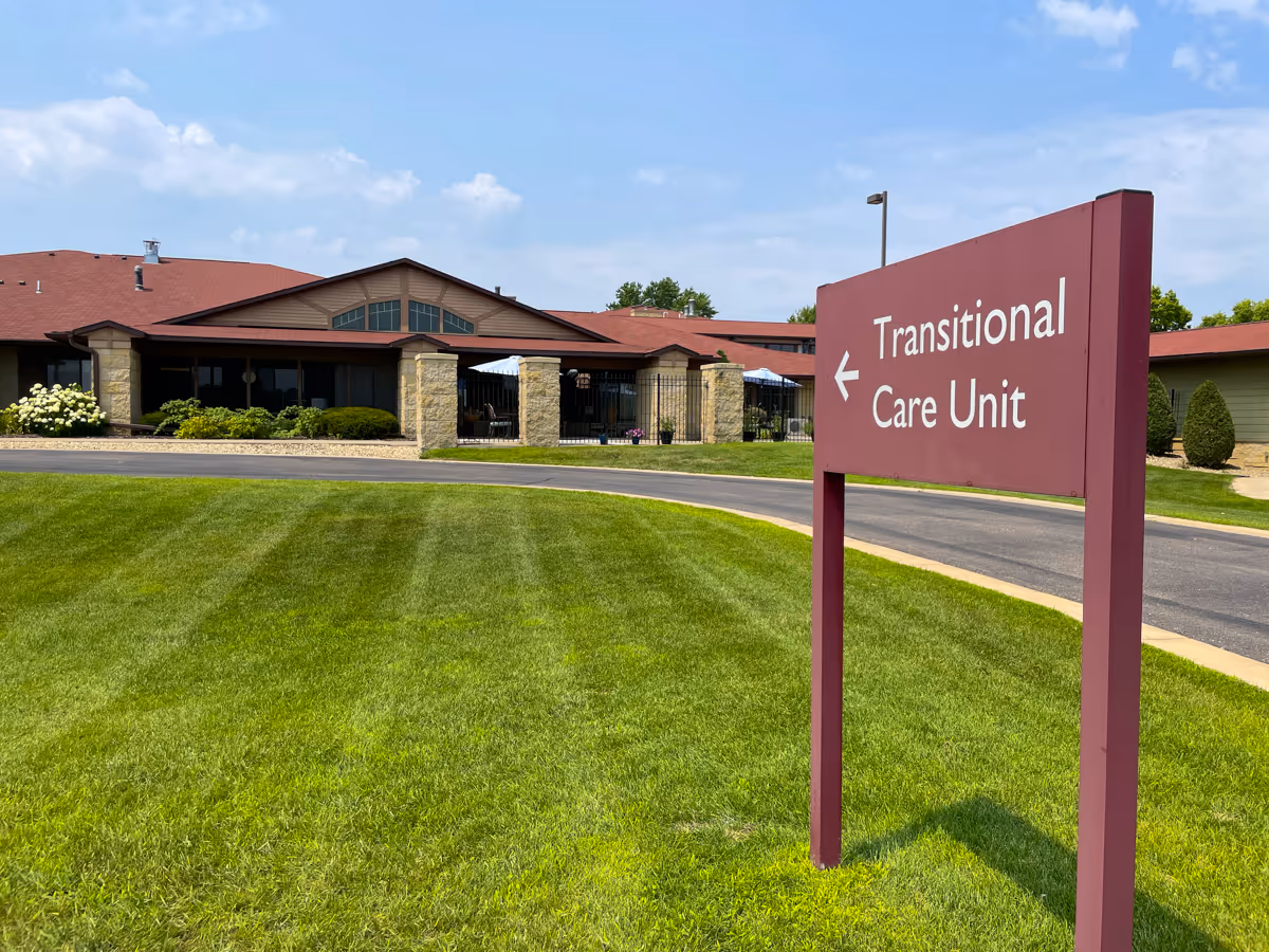 Outdoor view of a senior living facility with a well-maintained green lawn and a maroon sign pointing left that reads 'Transitional Care Unit'. The building has a red roof and stone pillars with a covered patio area visible.