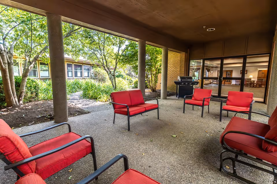 Covered outdoor patio area with red cushioned chairs arranged in a circle on a textured concrete floor. The patio is supported by round columns and overlooks a garden with trees and shrubs. A black grill is positioned near the glass doors leading inside the building.