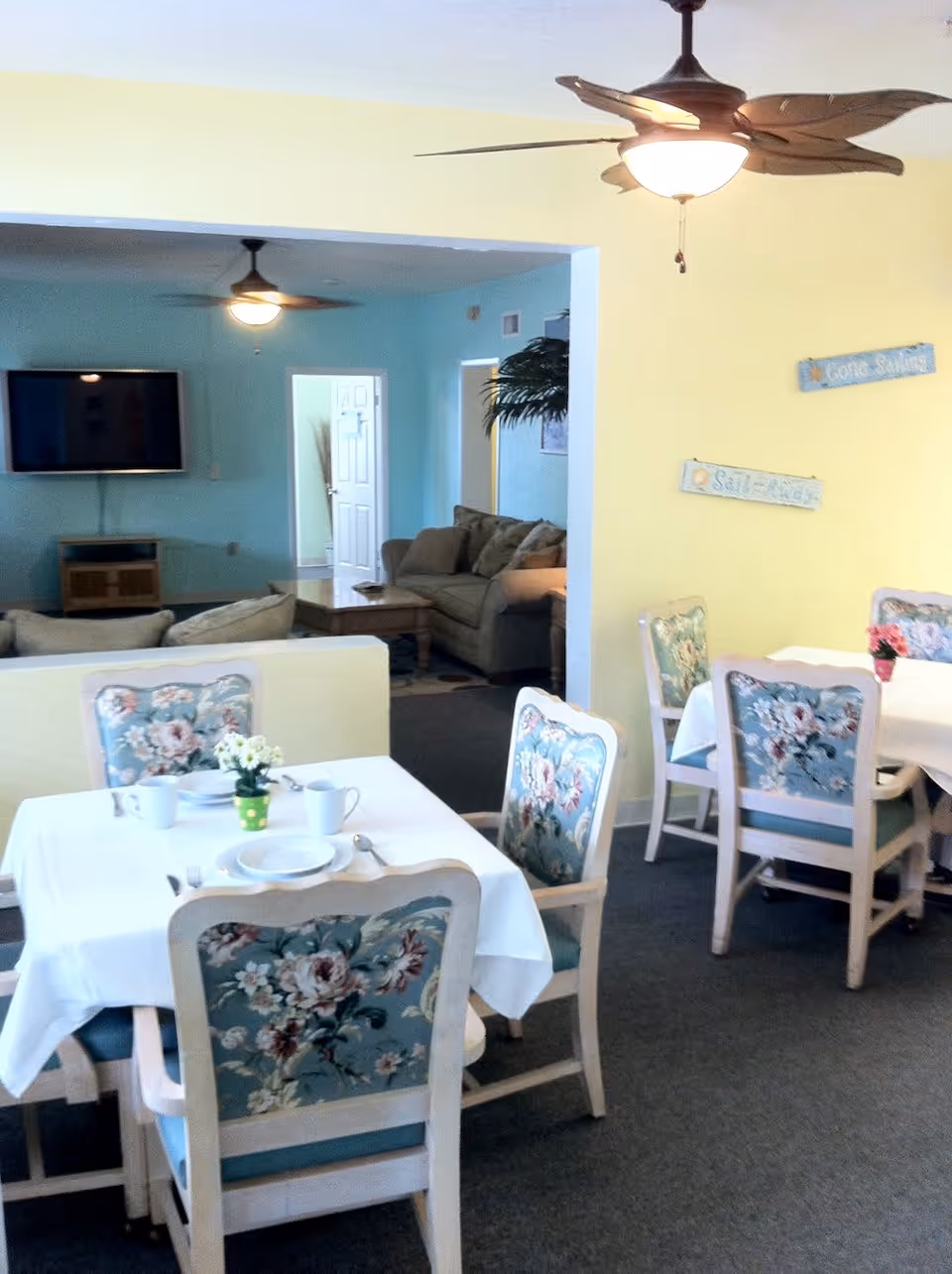 Dining room with white-clothed tables and floral-upholstered chairs, leading to a living area with couches and a wall-mounted TV.