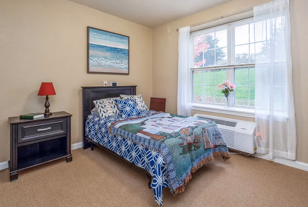 A cozy bedroom with a neatly made bed featuring blue and white patterned bedding and a decorative throw blanket. There is a dark wooden nightstand with a red lamp and a book on it. A large window with white curtains lets in natural light, and a vase with pink flowers sits on the windowsill. A painting of an ocean scene hangs on the beige wall above the bed.