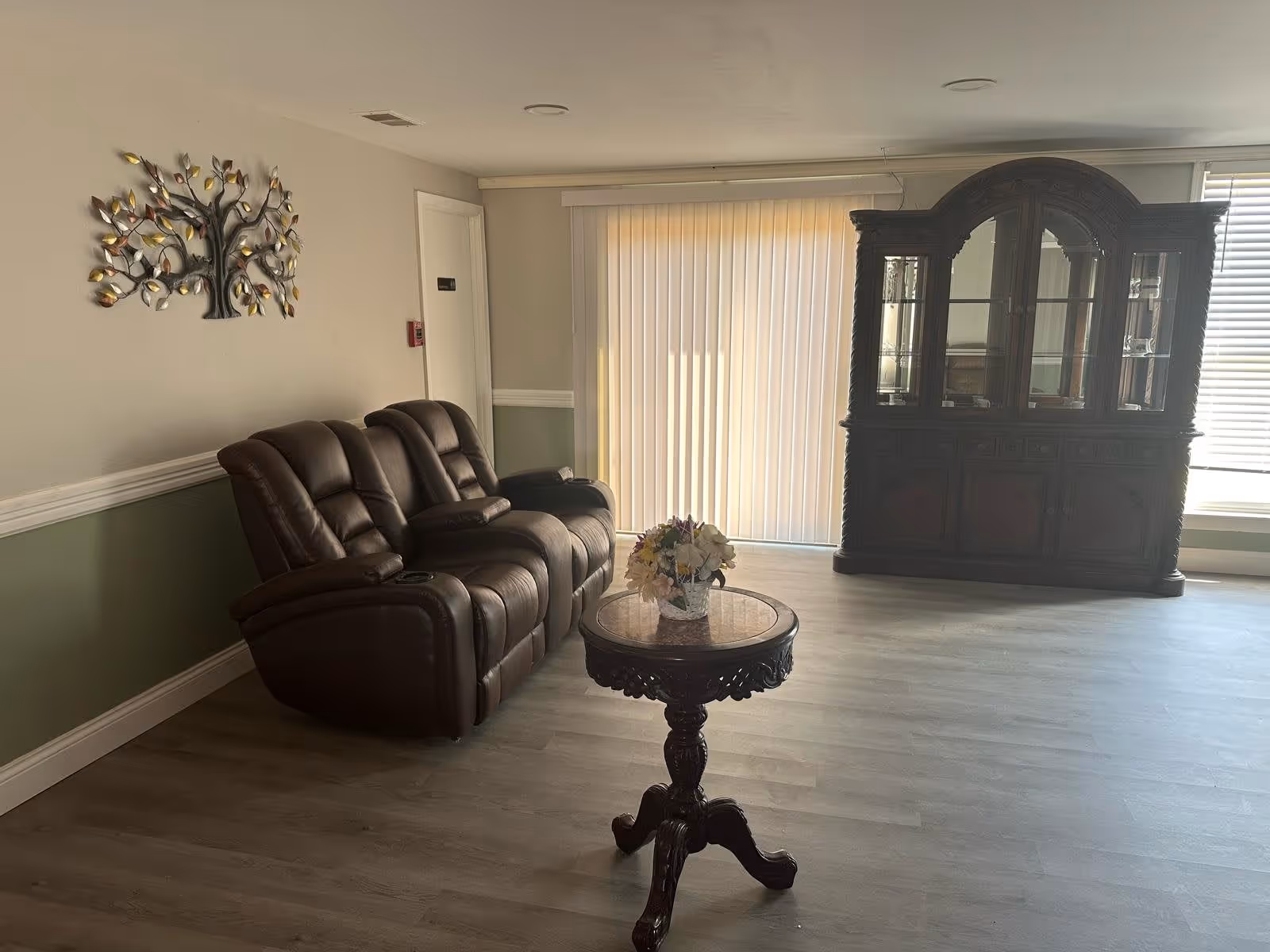 A living room with a brown leather reclining sofa against a two-tone wall with green and beige colors. A decorative metal tree wall art hangs above the sofa. In front of the sofa is a round wooden table with intricate carvings, holding a small flower arrangement. To the right, there is a large wooden china cabinet with glass doors. Vertical blinds cover a sliding glass door, and a window with horizontal blinds is partially visible.