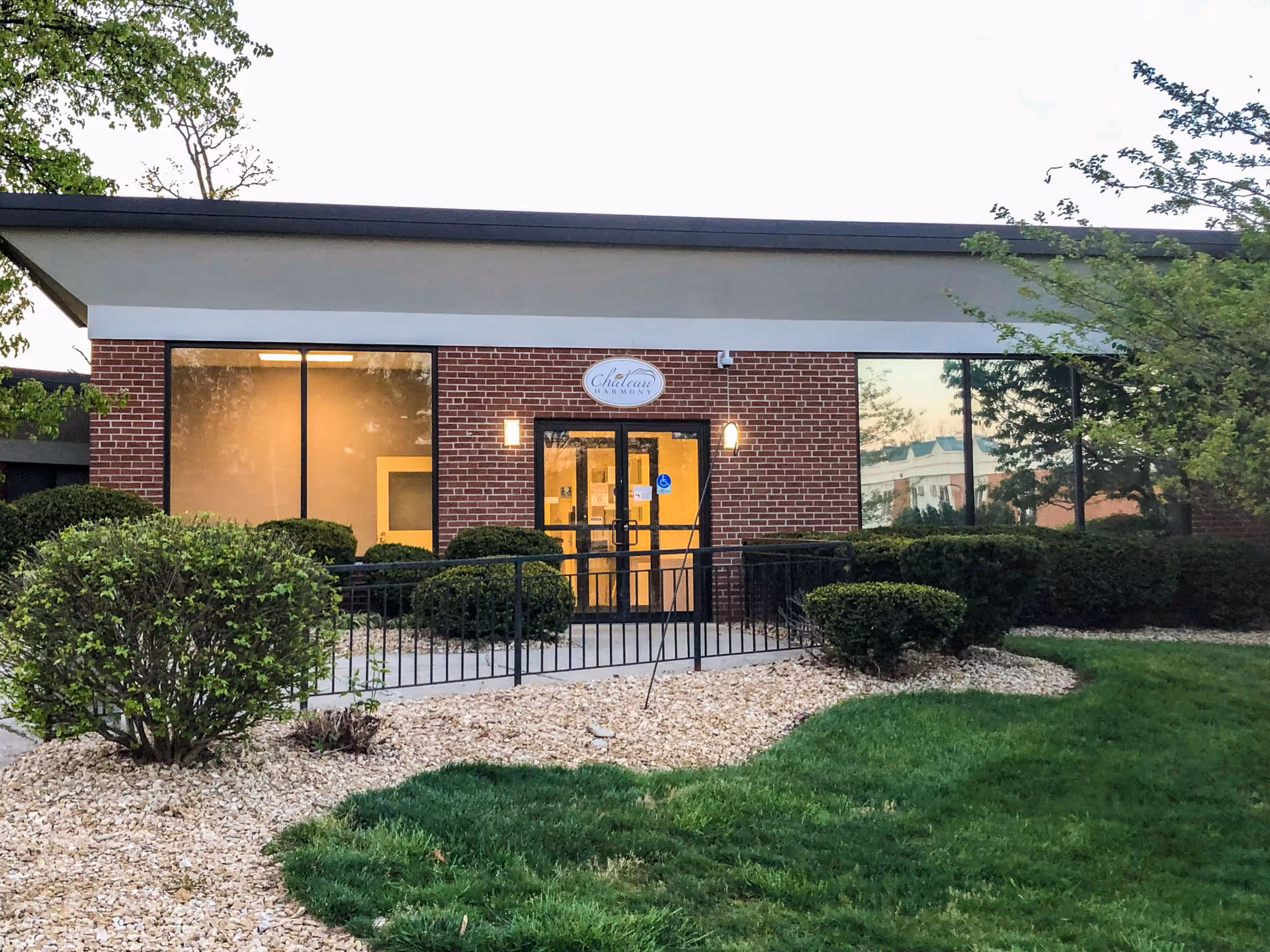 Exterior view of a single-story brick building with large windows and a glass door entrance. The building is surrounded by neatly trimmed bushes, a small rock garden, and a green lawn. There is a sign above the entrance that reads 'Chateau Harmony'.