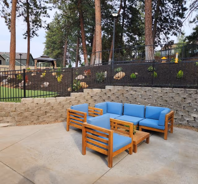 Outdoor seating area with wooden furniture featuring blue cushions arranged on a concrete patio. Behind the seating area is a retaining wall made of stone blocks, with a landscaped garden bed above it containing small plants and large trees. A black metal fence runs along the top of the retaining wall.