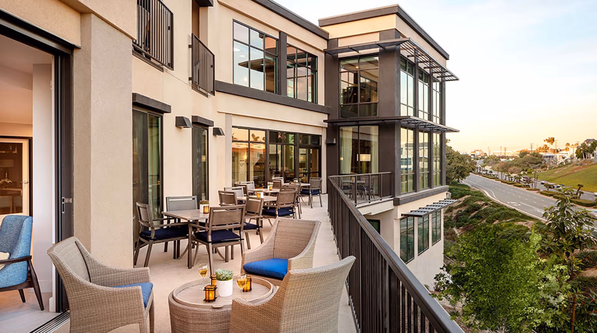 Outdoor balcony area of a senior living facility with several tables and chairs arranged for seating, overlooking a road and greenery. The building has large windows and a modern design.