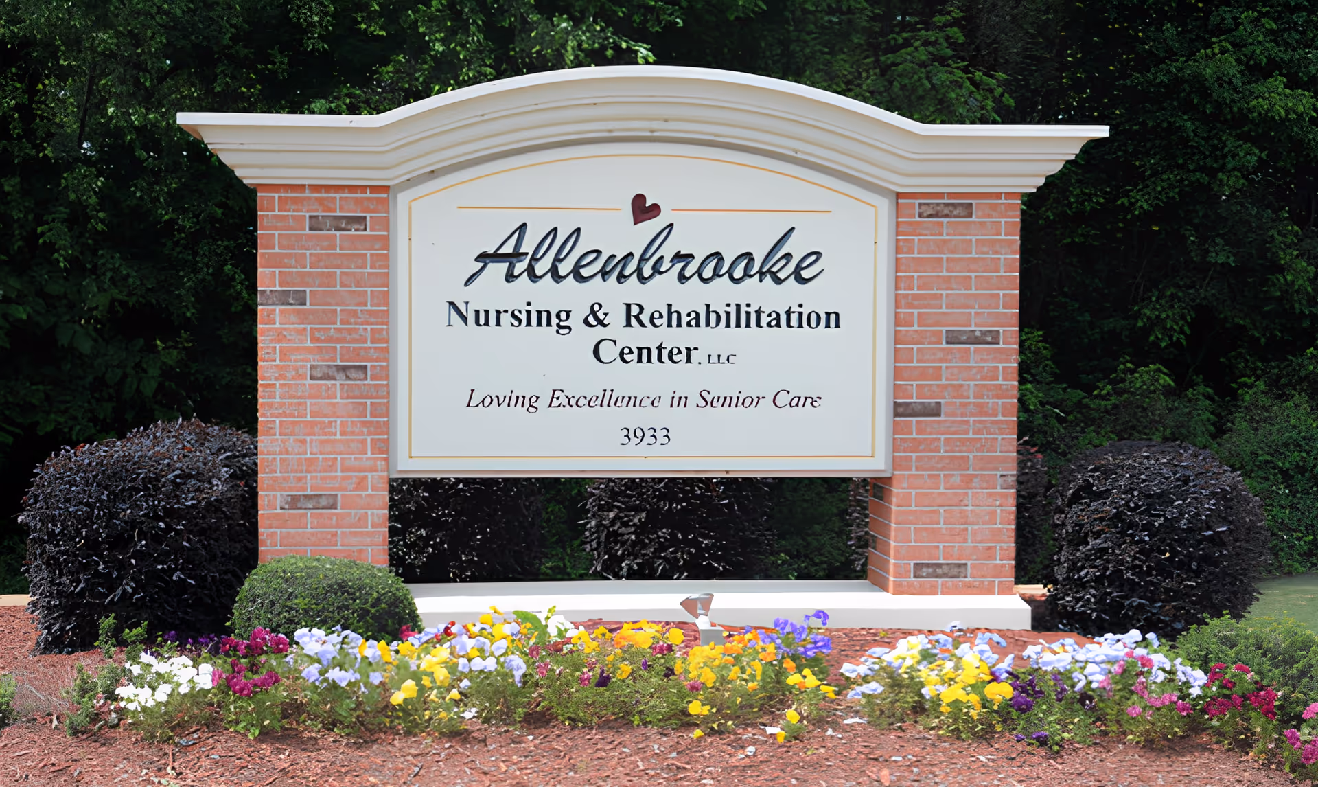 A large brick and white sign for Allenbrooke Nursing & Rehabilitation Center, LLC, surrounded by colorful flowers and green bushes with trees in the background.