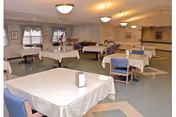 Spacious dining room with multiple tables covered in tablecloths, chairs, overhead lighting, and a small seating area in the back.