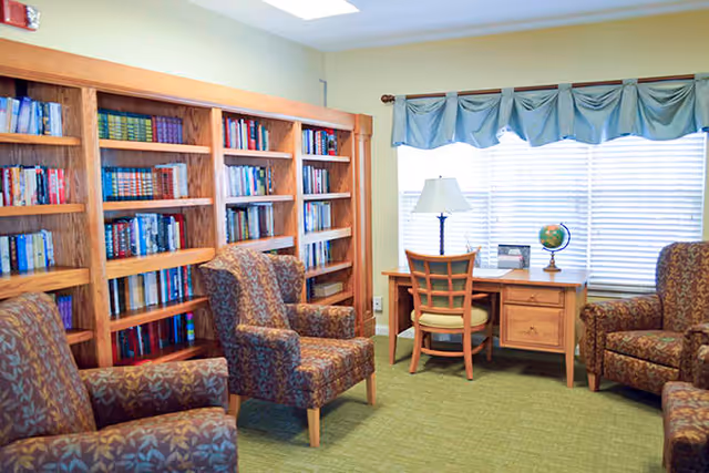 A cozy interior room with a green carpet, three patterned armchairs, a wooden desk with a chair, a table lamp, a globe, and a large bookshelf filled with books against the wall. A window with blinds and a blue valance is behind the desk.